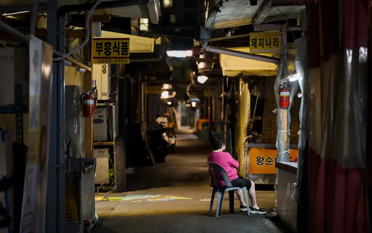 An elderly woman sits alone in a quiet alley market in Seoul, South Korea at night.