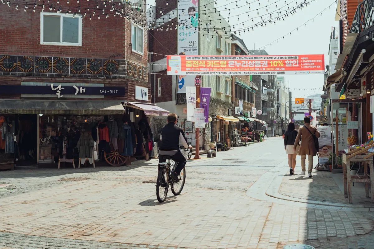 在首爾市的熱鬧街道上，有當地商店與腳踏車騎士，呈現韓國都市生活的精髓。